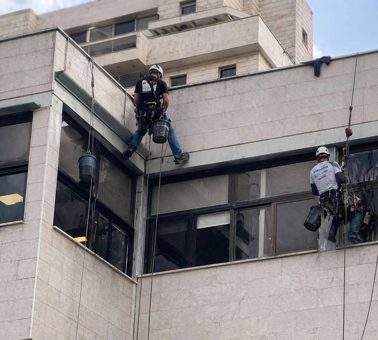 שני עובדים עם קסדות ורתמות תלויים בחבלים לצד חזית בניין